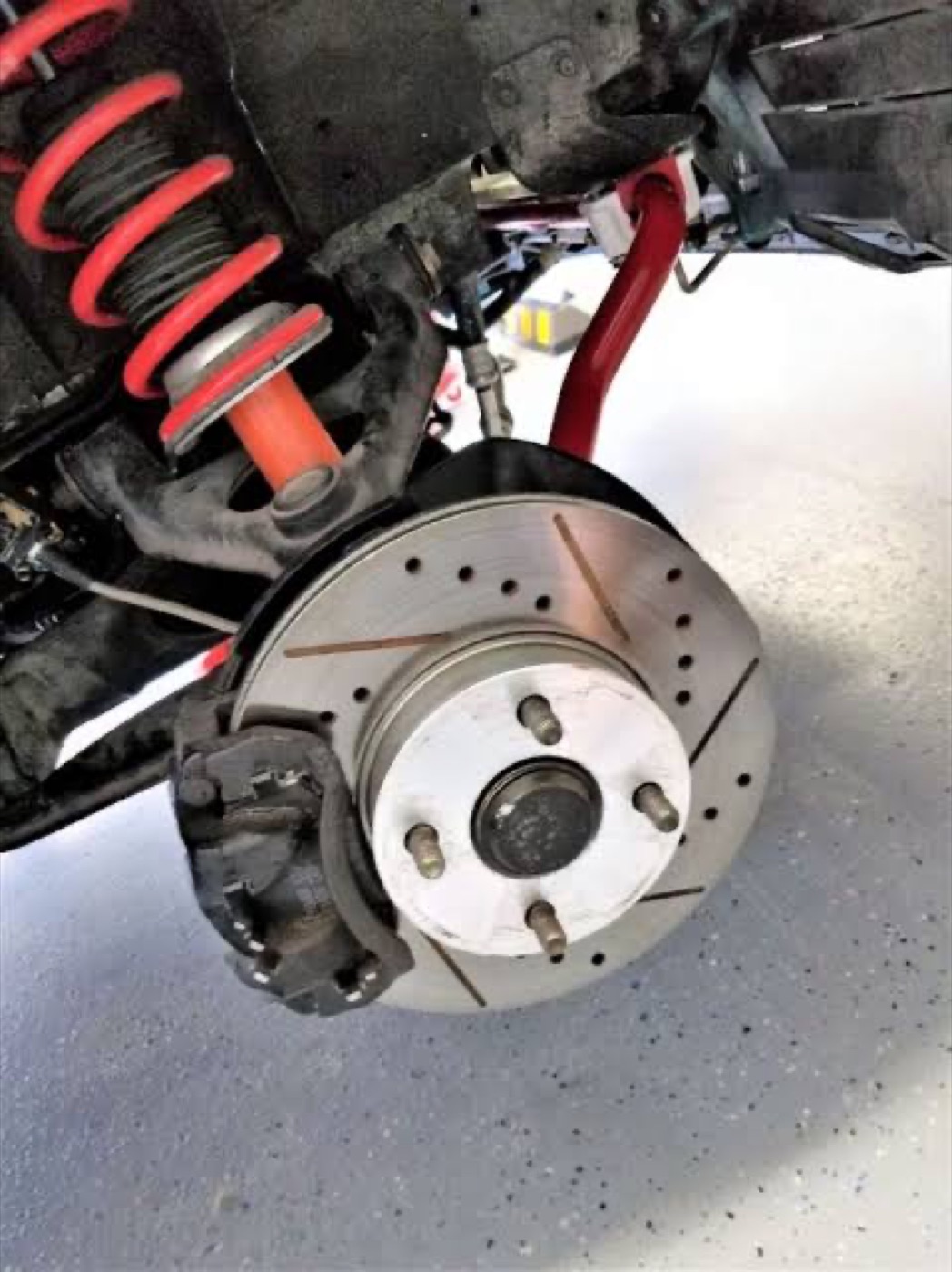A slotted and drilled brake rotor with the caliper off, coilover and sway bar visible